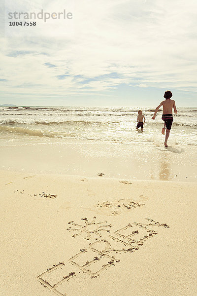 Spanien  Mallorca  Zwei kleine Jungen am Strand laufen ins Meer  Wort Ferien im Sand geschrieben