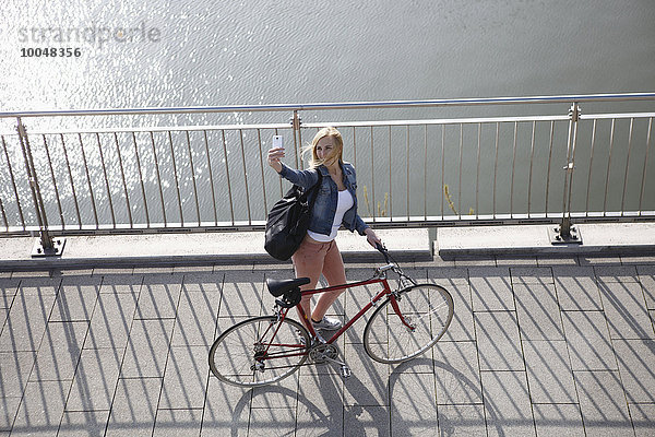 Deutschland  Düsseldorf  blonde Frau mit Fahrrad vor dem Rhein stehend mit einem Selfie mit Smartphone
