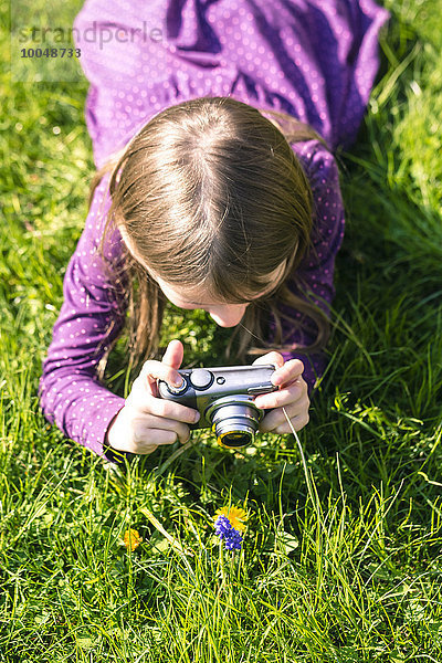 Kleines Mädchen  das auf der Wiese liegt und eine Blume fotografiert.