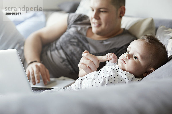 Junger Vater mit Laptop und Baby auf der Couch liegend