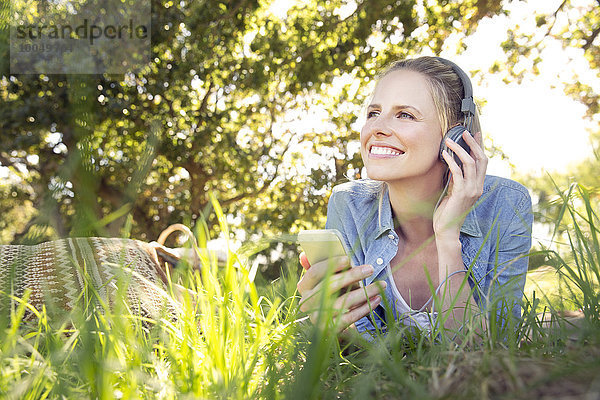Lächelnde Frau liegt auf der Wiese und hört Musik vom Smartphone.