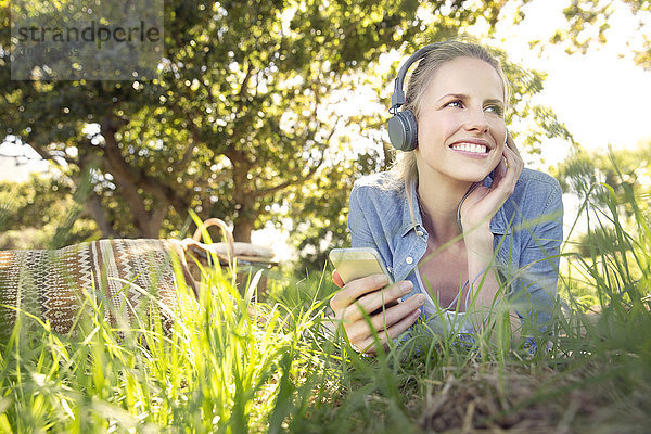 Lächelnde Frau liegt auf der Wiese und hört Musik vom Smartphone.
