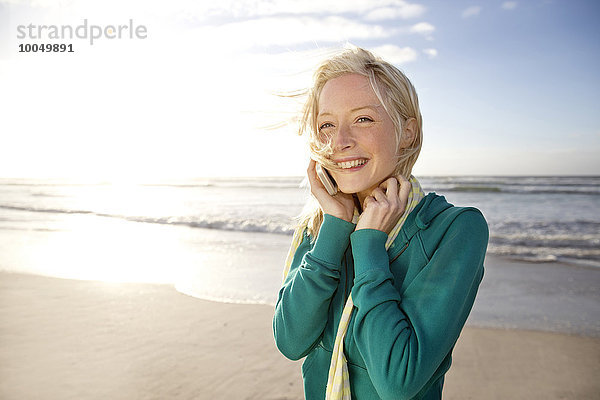Lächelnde junge Frau am Strand bei Sonnenaufgang per Handy