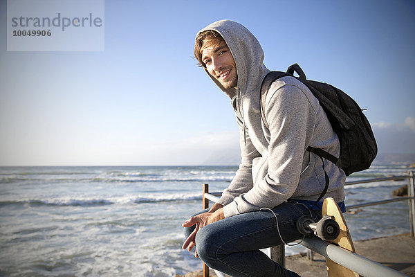 Lächelnder junger Mann sitzt auf einem Geländer an der Küste