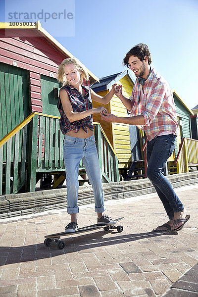 Junger Mann hilft Freundin auf Skateboard in bunten Strandhütten