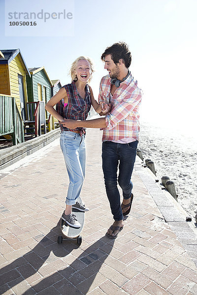 Junger Mann hilft Freundin auf Skateboard an der Strandpromenade