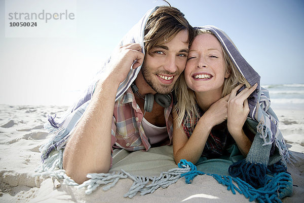 Ein glückliches junges Paar liegt am Strand unter einer Decke.