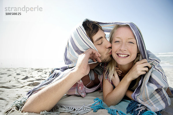 Fröhliches junges Paar am Strand liegend unter einer Decke küssend