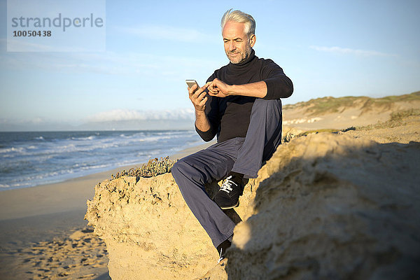 Südafrika  Porträt eines Mannes  der auf einem Felsen am Strand sitzt und ein Smartphone benutzt.