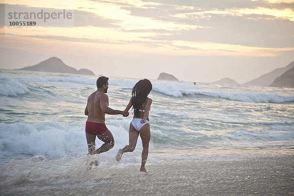 Mittleres erwachsenes Paar  das bei Flut am Strand entlang läuft  Hand in Hand  Rückansicht