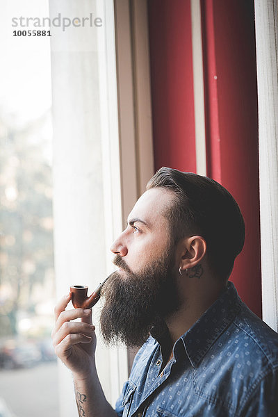 Junger bärtiger Mann raucht Pfeife am Fenster