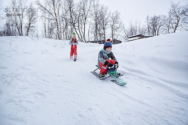 Zwei Brüder auf Schlitten und Schneescooter auf schneebedecktem Hügel  Hemavan  Schweden