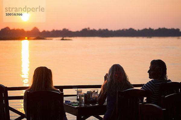 Rückansicht von drei erwachsenen Freunden  die den Sonnenuntergang über dem Mekong River  Don Det  Laos beobachten.