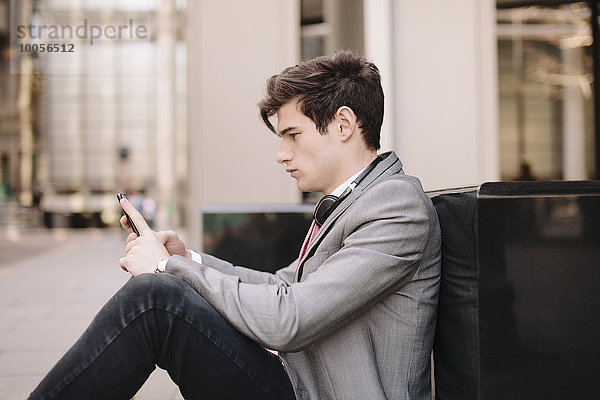 Ein junger Geschäftsmann sitzt auf dem Bürgersteig und liest Smartphone-Nachrichten.