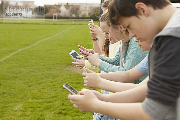 Reihe von fünf Jungen und Mädchen beim Lesen von Smartphone-Textnachrichten auf dem Fußballplatz