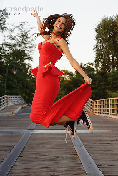 Porträt einer jungen Frau in einem schicken roten Kleid und Baseballschuhen  die in Bewegung springen.