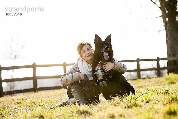 Porträt einer mittleren erwachsenen Frau  die mit ihrem Hund auf dem Feld sitzt.