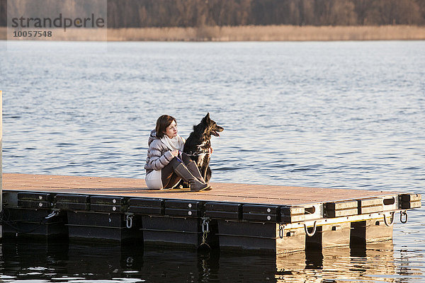 Mittlere erwachsene Frau und ihr Hund sitzen auf dem Pier am See.