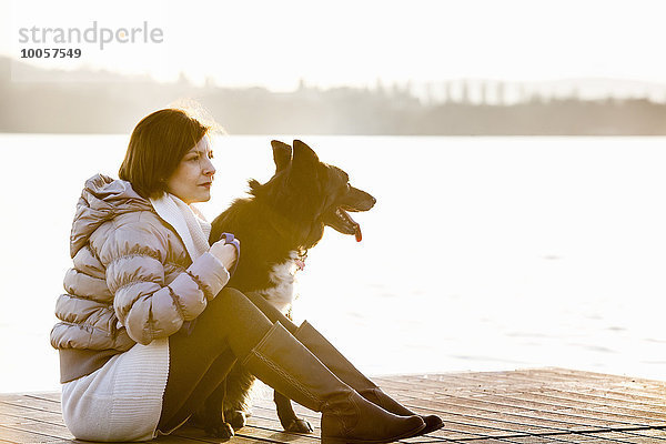 Mittlere erwachsene Frau und ihr Hund sitzen auf dem sonnendurchfluteten Pier am See.