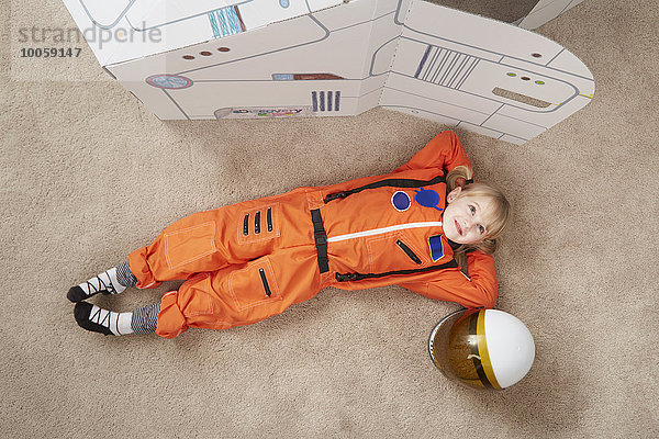 Junges Mädchen beim Spielen  im Astronauten-Outfit  neben einem Karton-Raumschiff liegend  erhöhte Ansicht
