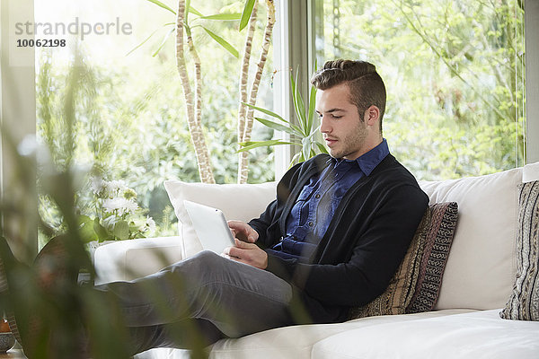 Mann entspannt sich auf dem Sofa mit den Füßen nach oben und schaut auf ein digitales Tablett.