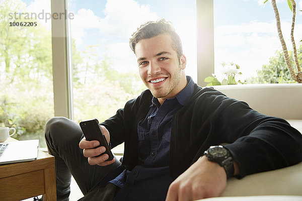 Porträt eines lächelnden Mannes  der auf dem Boden sitzt und ein Smartphone hält.