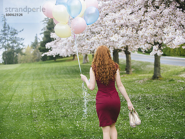 Rückansicht einer jungen Frau mit langen  gewellten roten Haaren und einem Haufen Luftballons  die im Frühlingspark spazieren gehen.