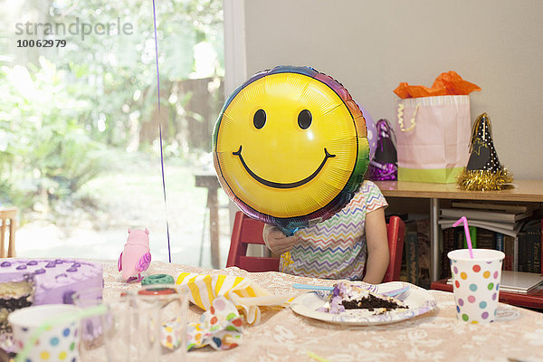 Mädchen sitzend am Geburtstagstisch mit Kuchenspiel mit Smiley-Gesichtsballon