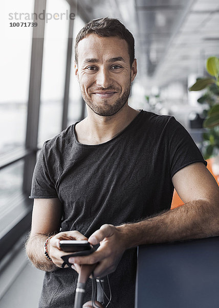 Porträt eines lächelnden Geschäftsmannes  der sein Handy in der Hand hält  während er sich auf die Kabine im Büro stützt.