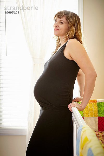Schwangere Frau  die sich im Kinderzimmer an die Krippe lehnt.