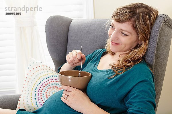 Schwangere Frau isst Suppentasse auf Wohnzimmersessel
