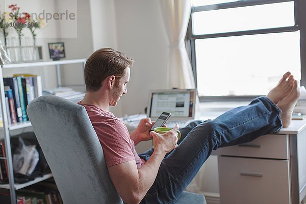 Mid Erwachsene Mann mit Füßen auf dem Schreibtisch lesen Smartphone-Texte