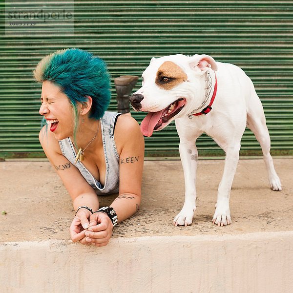 Junge Frau mit bunten Haaren  auf dem Boden neben dem Hund liegend  lachend