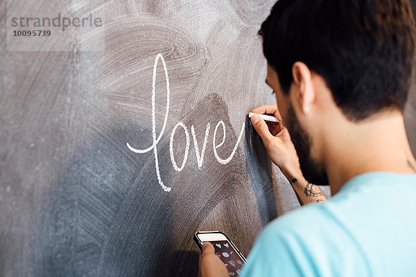 Junger Mann  das Wort Liebe auf die Tafel schreiben  Smartphone halten  Rückansicht