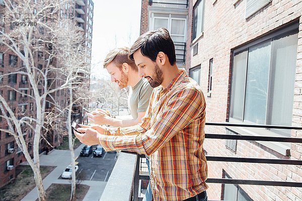 Männliches Paar auf dem Balkon stehend  mit Blick auf Smartphones