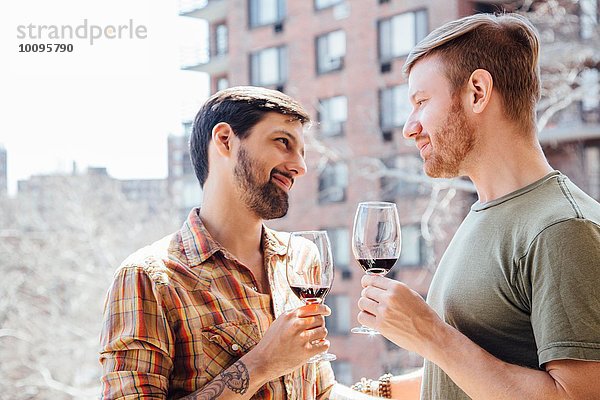 Männliches Paar steht auf dem Balkon  hält Weingläser  von Angesicht zu Angesicht