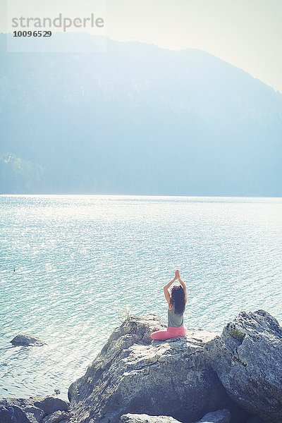 Mittlere erwachsene Frau  auf Felsen sitzend  am See  in Yogastellung  Rückansicht