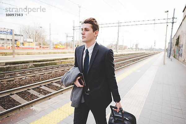 Junger Geschäftsmann  der mit dem Koffer am Bahnsteig entlanggeht.