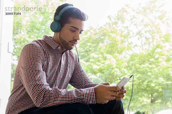 Junger Mann beim Auswählen von Musik auf dem Smartphone im Café-Fenstersitz