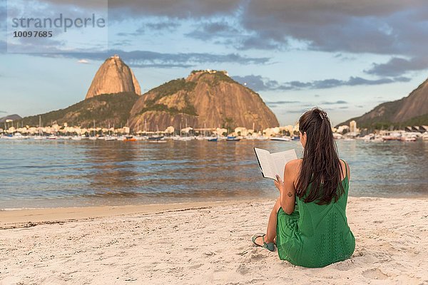 Mittlere erwachsene Frau am Strand sitzend  Lesebuch  Zuckerhut im Hintergrund  Botafogo-Bucht  Rio de Janeiro  Brasilien