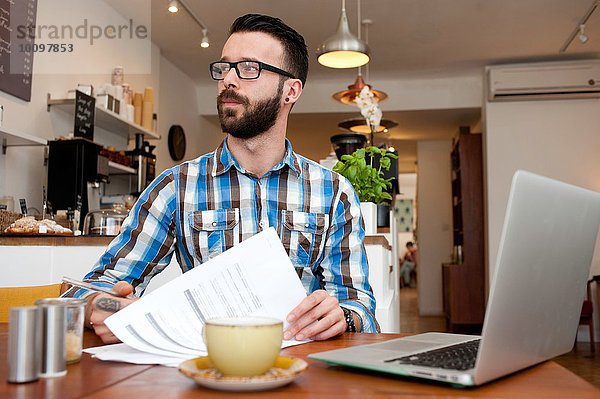 Männlicher Kunde liest Papierkram und benutzt den Laptop am Kaffeetisch.