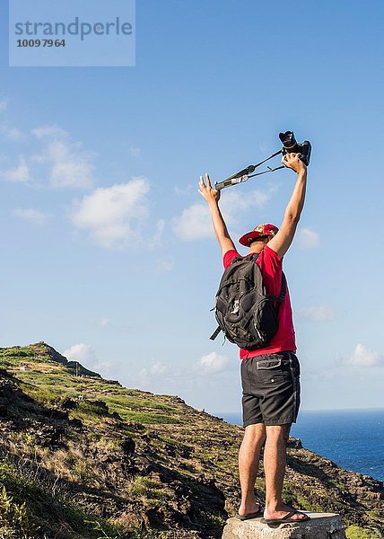Junger männlicher Tourist  der seine digitale Spiegelreflexkamera auf dem Küstenweg von Makapuuu  Oahu  Hawaii  USA  hochhält.