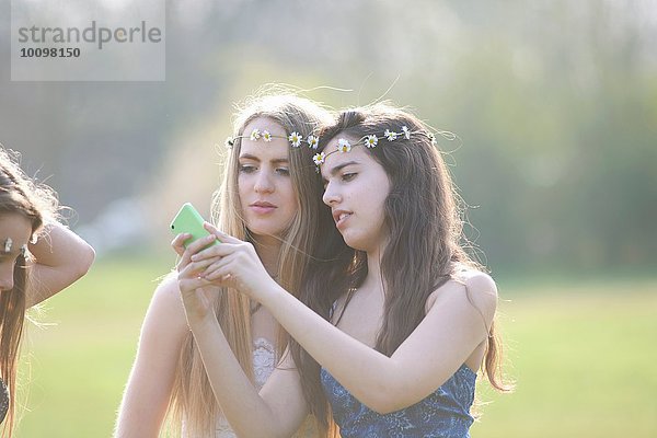 Zwei Teenager-Mädchen in Gänseblümchen-Kopfbedeckungen beim Lesen von Smartphone-Texten im Park