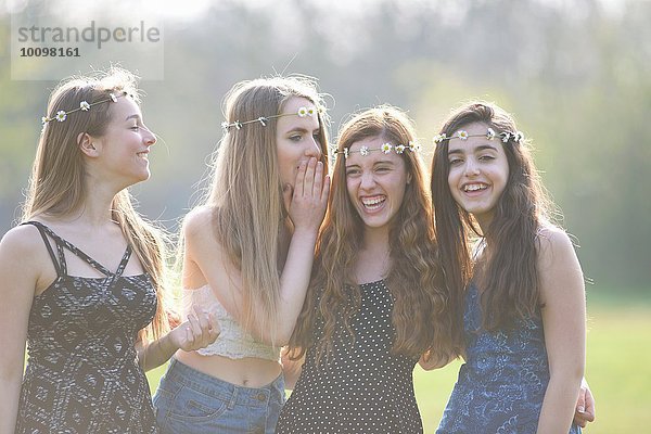 Vier Teenager-Mädchen in Gänseblümchen-Kopfbedeckungen kichern im Park.