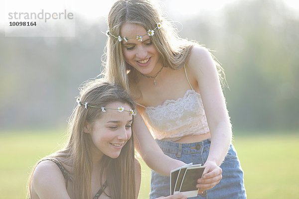 Zwei Teenager-Mädchen in Gänseblümchen-Kopfbedeckungen  die sich Sofortbilder im Park ansehen.