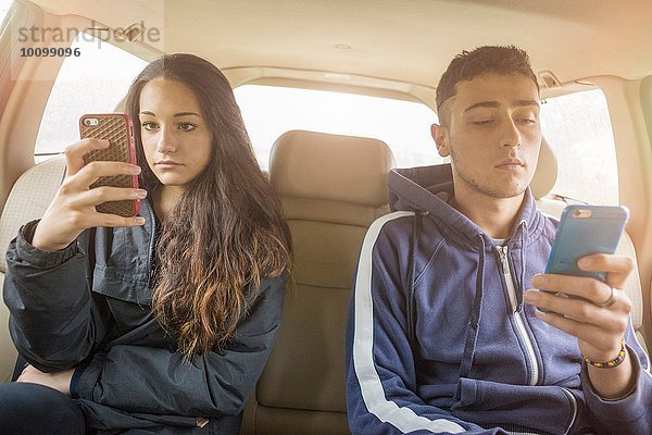 Teenager-Mädchen und junger Mann lesen separate Smartphone-Texte im Auto Rücksitz