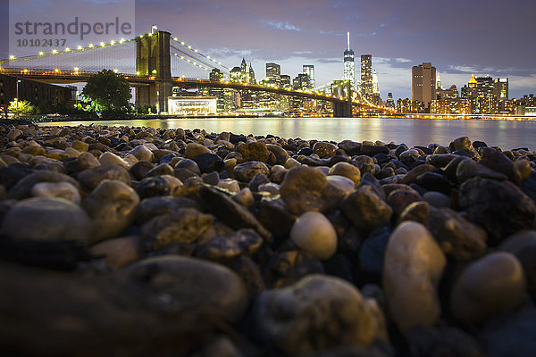 Nachtansicht in Richtung Manhattan von Brooklyn aus  mit der Brooklyn Bridge  die den East River überspannt  das felsige Ufer im Vordergrund.