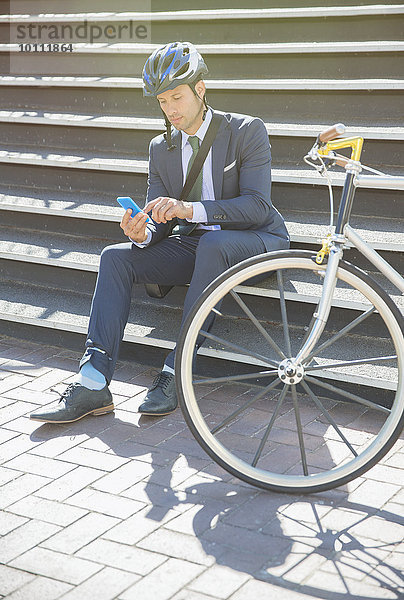 Geschäftsmann im Anzug und Helm SMS mit Handy neben dem Fahrrad auf der sonnigen Stadttreppe