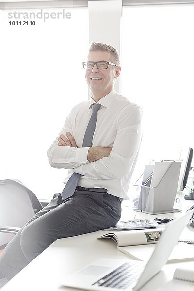 Portrait selbstbewusster Geschäftsmann mit Brille auf dem Schreibtisch sitzend mit gekreuzten Armen