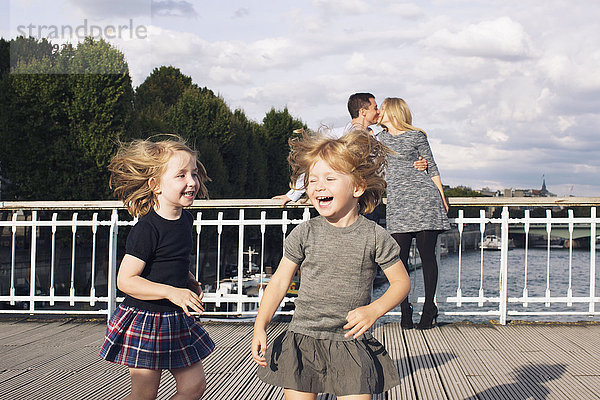 Kleine Mädchen spielen im Freien  während ihre Eltern sich im Hintergrund küssen.